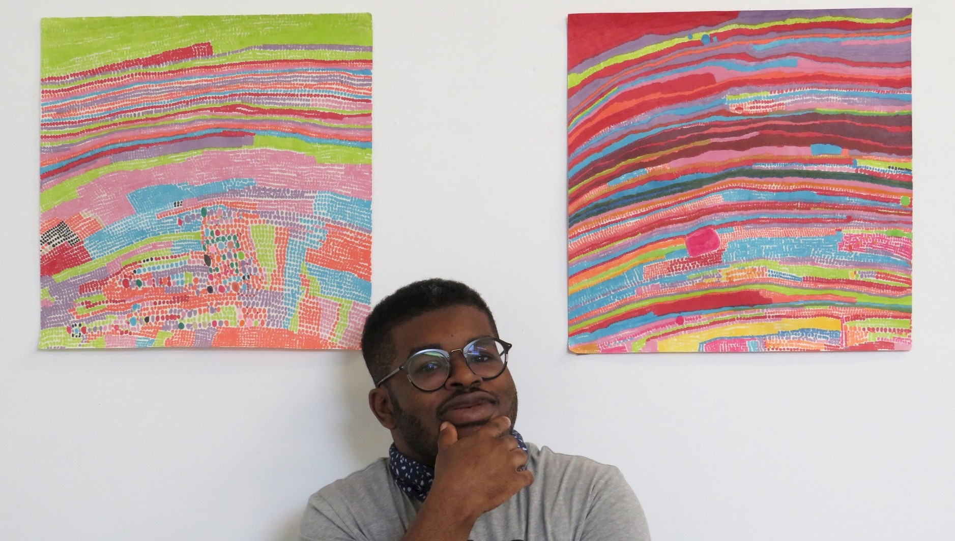 Artist Jorell Bonnick poses with his work. He is a young black man with a trim beard and moustache, wearing a neckerchief and glasses.